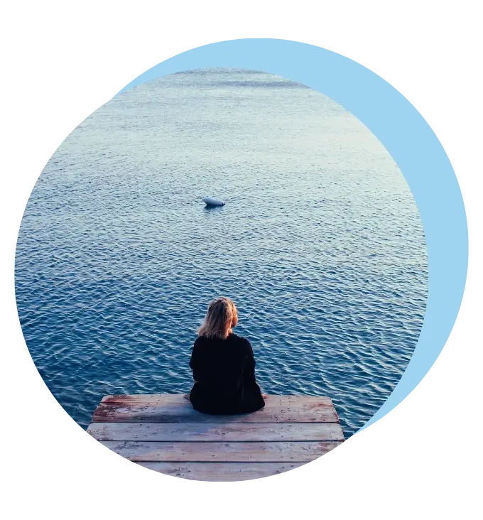 Kobieta siedząca tyłem, nad wodą, wpatrująca się w krajobraz